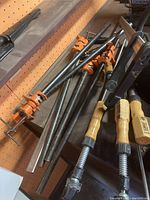 Grouped metal bar clamps on a workbench, showing metal bars and orange clamp heads with wooden handles.