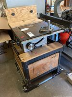 Full view of Craftsman table saw on wooden cabinet base with metal frame and wheels. Handmade tool accessory mounted on table. Bolts stored inside cabinet.