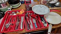 Overview of a large set of kitchen utensils spread across red trays with two frying pans on the side, one with glass lid and one without.