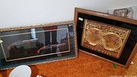 Two framed pieces on floor: left is flintlock pistols in shadow box frame; right is golden metallic double hemisphere world map art