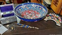 Wide view showing the large, detailed ceramic bowl with colorful floral and geometric patterns, the metal jewelry box with repousse decoration and purple velour lining, the necklace made of enameled metal beads in blues and silvers, and a portion of the wooden painted tribal figurines.
