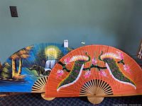 Two large hand painted fabric fans with wooden folding frames shown side by side against a blue wall. One fan features a waterfall nature scene and the other an orange fan with peacocks.