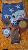 Lot laid out showing folded U.S. Department of State flag, three USS wooden plaques, British Police coin bank and two vintage lighters on an orange carpet.