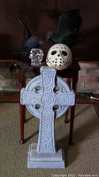 Photo showing large solid cast Celtic cross in front of table with skull candle holder, Jason mask, and two raven figurines on top.