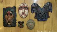 Five assorted masks and wall art arranged on wooden floor, showing variety in size, style, and material.