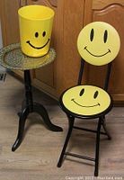 Yellow smiley face folding chair with black frame and matching yellow smiley face waste basket, both standing on the floor next to the occasional table.