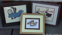 Three framed silk screen prints of indigenous art including a blue bird, two deer, and three owls grouped together on a dark surface.
