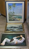 Photo of three original paintings arranged on floor: a beach scene with palm trees at top, rocky seascape in center, and figurative man falling at bottom.