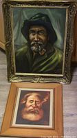Photo showing two framed paintings; one larger painting of a fisherman with pipe in ornate frame, and one smaller painting of old man with cigar in wood frame.