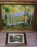 Two original paintings displayed together, a large birch tree acrylic painting in a gold frame and smaller rose cottage painting in a dark wood frame.