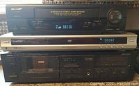 Front stacked view of Sharp VHS VCR on top, Toshiba DVD player in middle, Yamaha cassette deck on bottom, all powered on with digital displays active.