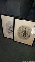 Full view of two wooden framed Dickens character prints leaning against a couch; both have mats, one has glass