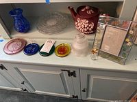 Photo of various glassware items including blue art glass vase, clear lunch plates, thumbprint plates in pink and blue, glass photo frame with pressed flowers, clear ribbed glass lamp shade, crystal pineapple, and ceramic teapot with floral design.