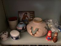 Shelved view of pottery collection including pottery bowl, covered trinket boxes, handpainted tile, and figurines.