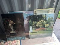 Photo of four framed museum posters leaning together, showing some titles and artwork, placed on a floor surface with natural light.