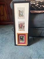 Photo shows three framed pieces leaning against couch and cabinet, two frames taller with three postcards each, one smaller frame with one postcard and Chinese child print.