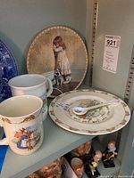 Photo showing two Bunnykins mugs, the Beatrix Potter spoon placed on a plate, and the "In Disgrace" decorative plate in the background.