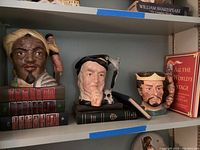 Three Royal Doulton character mugs of Shakespearean figures Macbeth, Hamlet, and Othello displayed on a shelf with several hardcover books behind them.