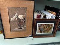Two framed Japanese woodblock prints displayed side-by-side on a shelf; left print showing female figure with parasol, right print showing busy market scene.