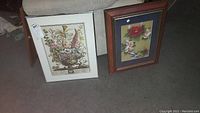 Both framed wall hangings shown side by side on a carpeted surface. The botanical print has a wooden frame, the Chinese silk painting has a metallic frame.