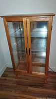 Front view of the wood display cabinet showing two glass paneled doors with black metal handles, glass on three sides, and wood finish.