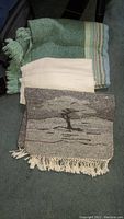 Three folded textiles displayed: green and blue wool cashmere shawl, natural off-white wool throw, and a woven African wall hanging with tree motif and fringes.