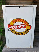 Full front view of the white metal popcorn stand showing colorful logo and surface rust on corners and edges.