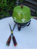 Side view of the vintage avocado green fondue pot with white floral designs, wooden handle, and lid with wooden knob. Six fondue forks with colored tips are laid out beside the pot.