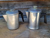 Pair of aluminum coffee pots with plastic handles and clear lid knobs, placed on wooden surface.