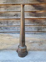 Front view of the antique brass fire hose nozzle standing upright on a wooden surface against a wooden wall background, showing the tapered cylindrical shape and base connection.