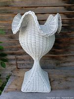 Front view of the white wicker plant stand showing the fluted petal-shaped top and bulbous central body increasing from a square base.