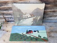 Both oil paintings displayed side by side, one canvas river scene painting and one folk art country scene painting on board.