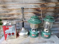 Front view of the two green Coleman lanterns, silver Coleman lamp, and red/white Coleman fuel can on wooden surface against wooden wall.