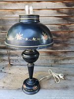 Full view of the black metal tole painted table lamp with floral designs, dome shade, and glass chimney on a weathered surface.