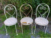 Three bent metal ice cream parlour chairs arranged outdoors on grass, two with white frames and one with rusty frame. All have Coca-Cola branded cushioned seats, one seat damaged.
