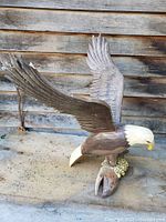 Side view of carved eagle perched on a fish, wing extended, showing wood texture and paint, slight damage visible on wing tip.