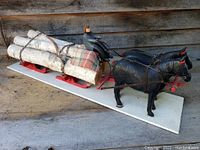 Side angle view of two black horses pulling red sleds loaded with birch logs and a blanket, with a painted wooden driver figure, mounted on a white base.