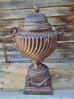 Full frontal view of urn with lid on, showing spiral ribbing on the body, ram head handles, and lid finial.