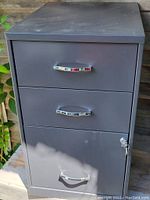 Front view of a gray metal 3 drawer filing cabinet showing the closed drawers and lock on the bottom drawer with key inserted.