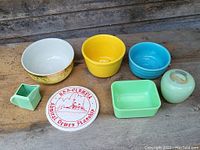 Seven colorful old pottery items including three bowls, a rectangular dish, a small container, a small vase, and a collectible ceramic plate all arranged on a wooden surface.