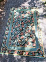 Full view of rectangular wool rug with a floral pattern and green borders placed outdoors on a gravel surface.