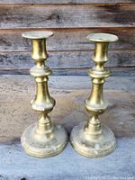 Front view of both brass Victorian candlesticks standing upright on a rustic surface against a wooden backdrop, showing their size and shape.