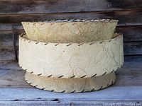 Photo of the triple decker fibre lamp shade showing its three-tiered design, beige color, textured fibre material, and stitching around the edges, placed against a wooden background.