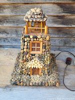 Front view of the handmade lighthouse lamp showing its detailed stone and wood construction, seashell accents on the roof and door, and the power cord visible on the right side.