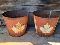 Pair of rusted vintage sap pails with white maple leaf emblem shown from the front, both set against a wooden backdrop.