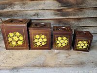 Four wooden canisters in descending size lined up, showing daisy floral artwork on front and lids with circular cutout handles