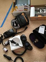 Overview of lot contents including Tasco binoculars with case, Nikon 35mm film camera, Kodak digital frame box, camera carrying case, film rolls, and chargers.