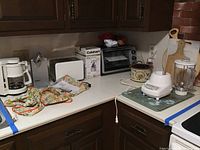 Photo of various kitchen appliances and items on counter including a white Krups coffee maker, Bella toaster, Cuisinart can opener and chopper, Black & Decker toaster oven, Proctor Silex blender, Rival crock pot, and assortment of cutting boards.