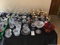 Wide view of collection including several clear crystal glasses, gold-rimmed goblets, and a red footed bowl arranged on a black cloth on table.