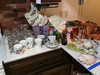 Wide view of the kitchen counter showing all the glassware, cups, colored vases, copper cookware, and kitchen textiles included in the lot.